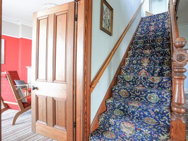 A wooden staircase with a patterned carpet and a wooden door next to a room with red walls and chairs at Llys Owen in Criccieth