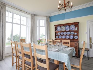 A dining room with a wooden table and chairs set with plates and glasses next to large windows and a wooden cabinet with blue and white plates at Llys Owen in Criccieth