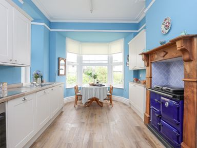 A kitchen with white cabinets a wooden floor a dining table by a bay window and a blue stove at Llys Owen in Criccieth