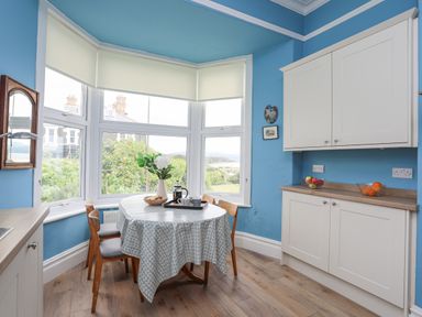 A kitchen dining area with a round table and chairs near large windows with white cabinets on the side at Llys Owen in Criccieth