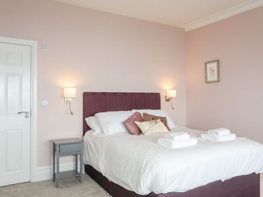 A bedroom with a double bed with white bedding and pillows two wall lamps a small grey bedside table and a framed picture on the wall at Llys Owen in Criccieth