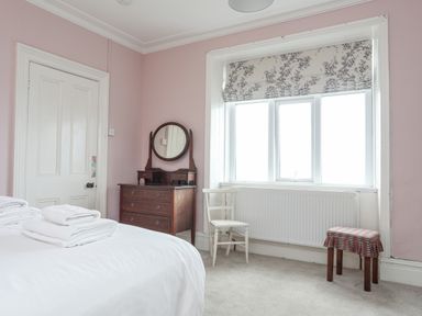 A bedroom with a bed and white towels a wooden dresser with a round mirror a white chair and a small stool by the window at Llys Owen in Criccieth