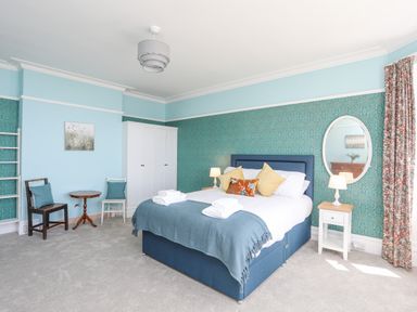 A bedroom with a blue upholstered bed with pillows and folded towels by a patterned wallpapered wall with a white wardrobe two chairs a small round wooden table bedside tables with lamps and a round mirror at Llys Owen in Criccieth