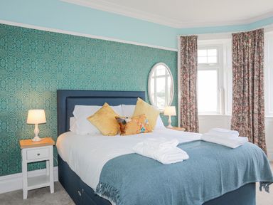 A bedroom with a bed and pillows lamps on bedside tables patterned curtains and a round mirror at Llys Owen in Criccieth