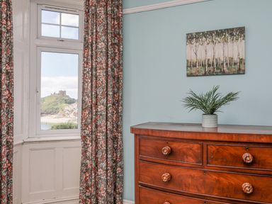A wooden dresser with a plant on top beside a window with floral curtains and a painting on the wall at Llys Owen in Criccieth