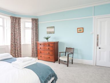 A bedroom with a bed with towels and blue blanket a wooden chest of drawers a chair and floral curtains at Llys Owen in Criccieth