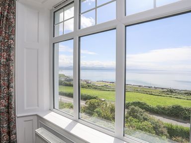 A large window with white frames showing a view of the sea green fields and a road at Llys Owen in Criccieth