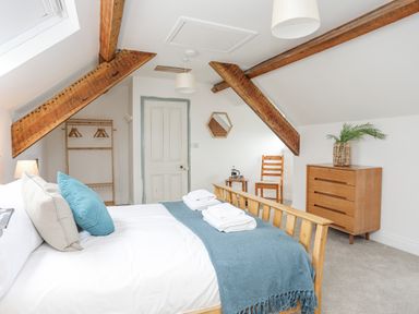 A bedroom with a wooden bed frame white bedding blue throw pillows and towels a wooden dresser chair and hanging rack at Llys Owen in Criccieth