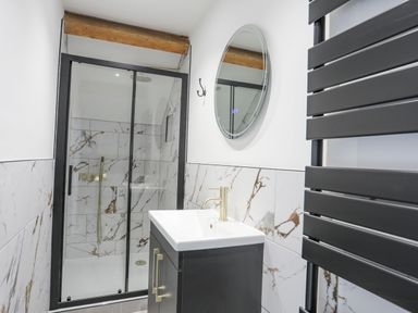 A bathroom with a glass shower enclosure a black vanity with a sink a round mirror and a black towel radiator at Llys Owen in Criccieth