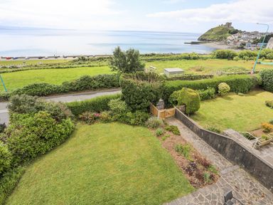 A view of a garden with bushes and a pathway overlooking a road green fields the sea and a hill with a castle Llys Owen Criccieth