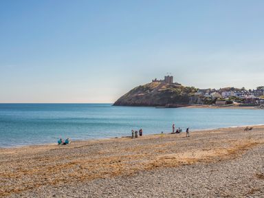 A pebble beach with people near the water and a hill with a castle and houses in the background at Llys Owen in Criccieth