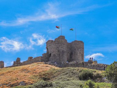 A stone castle ruin on a hill with two Welsh flags flying at Llys Owen in Criccieth