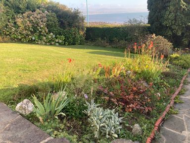 A garden with a flower bed next to a lawn and a view of the sea at Llys Owen in Criccieth