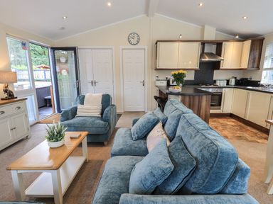 A living room with blue sofa and armchair a wooden coffee table and an open door leading outside at Hill Top Lodge in Bowness 65 White Cross Bay