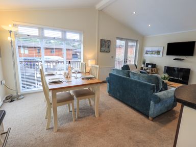 A dining table set for four beside large windows and a living room with blue sofas and a wall mounted TV at Hill Top Lodge in Bowness 65 White Cross Bay
