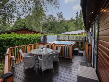 An outdoor wooden deck with a round glass table and four wicker chairs next to a hot tub and surrounded by greenery at Hill Top Lodge Bowness 65 White Cross Bay