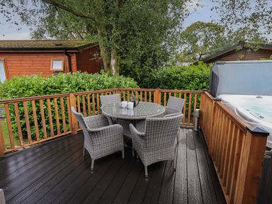 An outdoor deck with a round glass table and four wicker chairs next to a wooden railing and a hot tub at Hill Top Lodge in Bowness 65 White Cross Bay