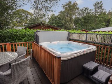 An outdoor deck with a hot tub with a cover and wicker chairs around a round glass table at Hill Top Lodge in Bowness 65 White Cross Bay