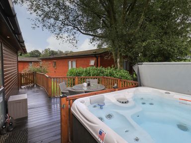 An outdoor deck with a hot tub and a round table with chairs near wooden cabins at Hill Top Lodge Bowness 65 White Cross Bay