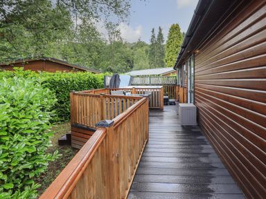 A wooden deck with railings and outdoor seating next to a wooden cabin surrounded by trees and bushes at Hill Top Lodge in Bowness 65 White Cross Bay