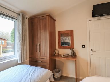 A bedroom corner with a wooden wardrobe, small desk with decorative items, wall mirror, window with curtain, and a door with a TV above at Hill Top Lodge in Bowness 65 White Cross Bay
