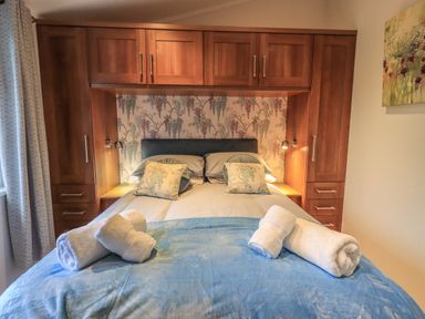 A bedroom with a double bed with floral pillows wooden cupboards curtains and rolled towels on a blue blanket at Hill Top Lodge in Bowness 65 White Cross Bay