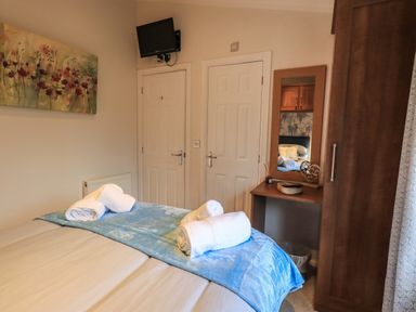 A bedroom with a bed topped with towels a wall-mounted televised two white doors a dressing table with a mirror and a wooden wardrobe at Hill Top Lodge in Bowness 65 White Cross Bay