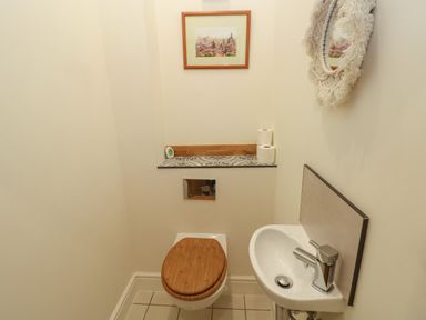 A small bathroom with a toilet with a wooden seat a wall-mounted sink a framed picture a round mirror and toilet paper rolls on a tiled shelf at Rudbaxton Water Rudbaxton near Haverfordwest
