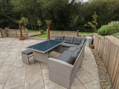 An outdoor patio with a gray wicker sectional sofa and glass-top table surrounded by trees at Rudbaxton Water in Rudbaxton near Haverfordwest