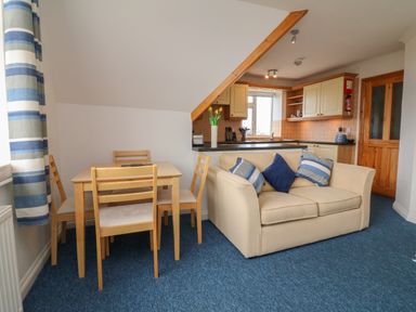 A small dining area with wooden table and chairs next to a beige sofa with blue cushions in an open plan kitchen and living space at 5 Seymour Villas in Woolacombe