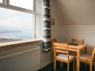 A dining area with a wooden table and four chairs near a large window with striped curtains at 5 Seymour Villas in Woolacombe