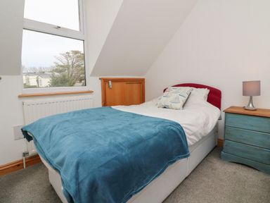A bedroom with a single bed covered with a blue blanket and a pillow with boat designs a blue bedside table with a lamp and a window with a view at 5 Seymour Villas in Woolacombe