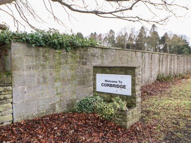 A stone wall with a welcome to Corbridge sign and some greenery at Willows in Corbridge
