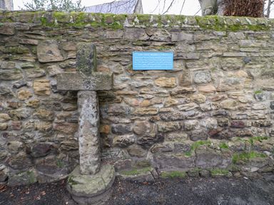 A stone cross mounted on a stone wall with a blue plaque explaining its history at Willows in Corbridge