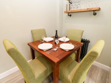 A small dining area with a wooden table set for four with green upholstered chairs and a wall shelf with glasses at Willows in Corbridge