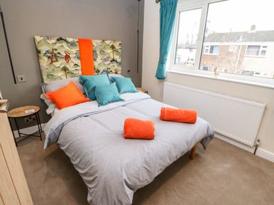 A bedroom with a double bed covered with gray bedding and teal and orange cushions next to a window with teal curtains at Willows in Corbridge