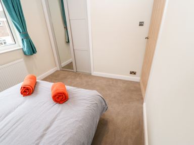 A bedroom with a bed covered by a gray sheet and two rolled orange towels a window with teal curtains a carpeted floor and a mirrored closet door at Willows in Corbridge