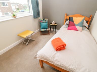 A bedroom with a single wooden bed white bedding with a colorful pillow and orange towel a small chair with floral and teal cushions a yellow stool and a small round black side table with tissues on it at Willows in Corbridge