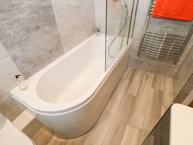 A bathroom with a white bathtub a glass shower screen a soap dispenser and a heated towel rail with an orange towel at Willows in Corbridge