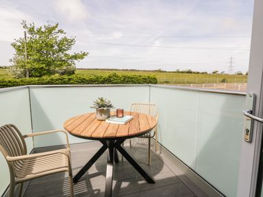A small balcony with a round wooden table, two beige chairs, a potted plant, a book, and a candle at The Hayloft in Llanfechell