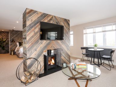 A living area with a wood-burning stove under a mounted TV a glass coffee table with books and a plant and a small dining table with two chairs near windows at The Hayloft in Llanfechell
