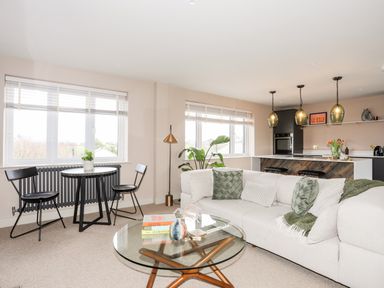 An open living room and kitchen with a round glass coffee table a white sectional sofa with green pillows a small black dining table with two black chairs and three pendant lights at The Hayloft in Llanfechell