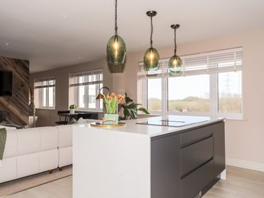 A kitchen island with pendant lights and flowers with a living area and windows in the background at The Hayloft in Llanfechell
