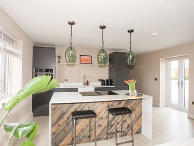 A kitchen with a wooden island and two stools three hanging green glass pendant lights and a vase of flowers at The Hayloft in Llanfechell