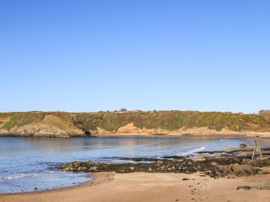 A beach with rocks and calm water under a clear sky with a green hillside in the background at The Hayloft in Llanfechell