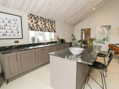 A kitchen with a black countertop island with stools and cabinets under a wooden ceiling at Garden House in Llanfechell