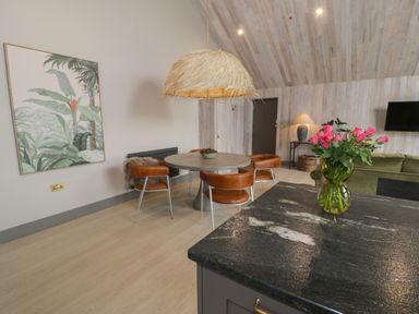 A dining area with a round table and four brown chairs a large plant painting and a kitchen countertop with pink flowers at Garden House in Llanfechell
