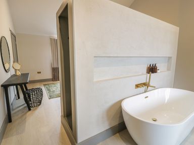 A bathroom with a white freestanding tub and gold faucet next to a wall with built-in shelf holding bottles and a room with a table mirror stool and rug at Garden House in Llanfechell
