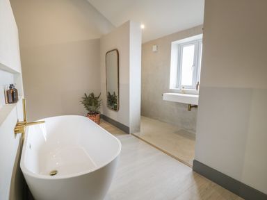 A bathroom with a freestanding bathtub gold faucet a full length mirror a potted plant and a floating sink under a window at Garden House in Llanfechell