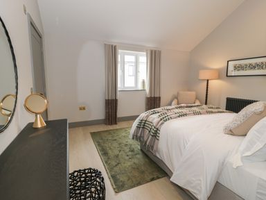 A bedroom with a bed covered by a plaid blanket a green rug next to the bed an armchair a floor lamp a framed picture on the wall and a black dresser with a mirror and a small round mirror at Garden House in Llanfechell